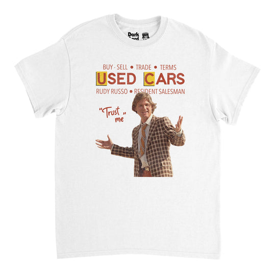 A vintage, 70s style white colored short sleeved t-shirt that says "Buy, Sell, Trade, Terms" above the title of iconic 1980 satirical film "Used Cars" and "Rudy Russo, Resident Salesman" above an image of Russo himself - portrayed by famous actor Kurt Russell - wearing a checkered brown suit and tie next to his popular quote, 'Trust me'.