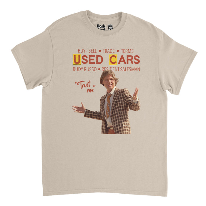A retro, sand colored 80s style souvenir t-shirt that says "Buy, Sell, Trade, Terms," "Used Cars,"  "Rudy Russo, Resident Salesman" and the quote "Trust Me" beside an image of character Rudy Russo wearing a checkered brown suit and tie, played by iconic famous actor Kurt Russell in the satirical comedy film "Used Cars" also starring Jack Warden as Luke Fuchs.