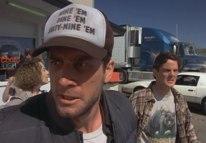 The movie character Sea Bass, played by Cam Neely, is in a parking lot with a mad expression, another man in a flannel and "Beer?" shirt with a bear behind him. He's wearing a trucker hat that says "Wine 'Em, Dine 'Em, Sixty-Nine 'Em" and is from the famous comedic film "Dumb and Dumber", which stars actors Jim Carrey and Jeff Daniels.
